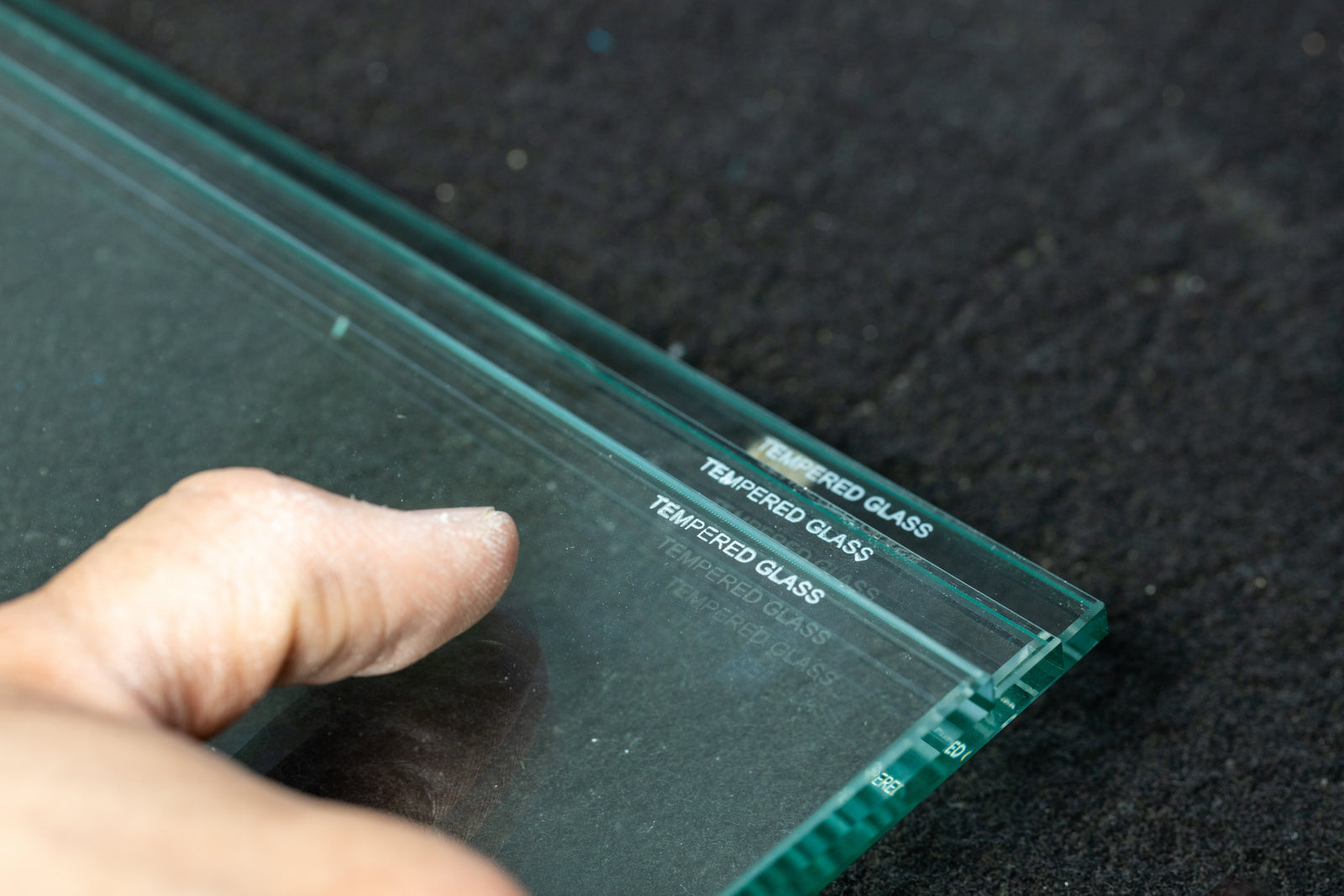 A close-up of a hand holding a clear tempered glass panel with white printed text reading 'TEMPERED GLASS' on a dark textured surface.
